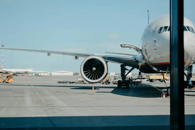 A large white commercial airplane parked on an airport tarmac with its large jet engine visible in the foreground and ground support vehicles nearby.